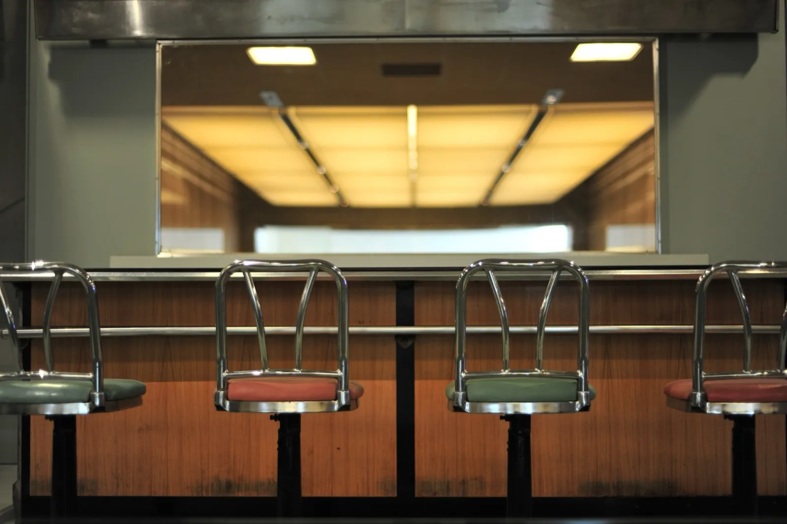 Four chrome stools with green and red vinyl seats in front of a wood-paneled lunch counter, under a stainless steel hood