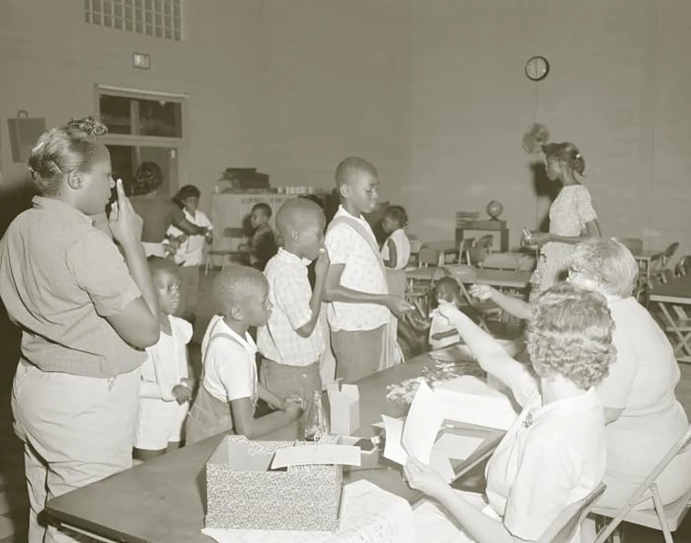Boys stand in line at a folding table where women sort paperwork, and one boy lifts a sugar cube to his mouth, in a school room in Georgia