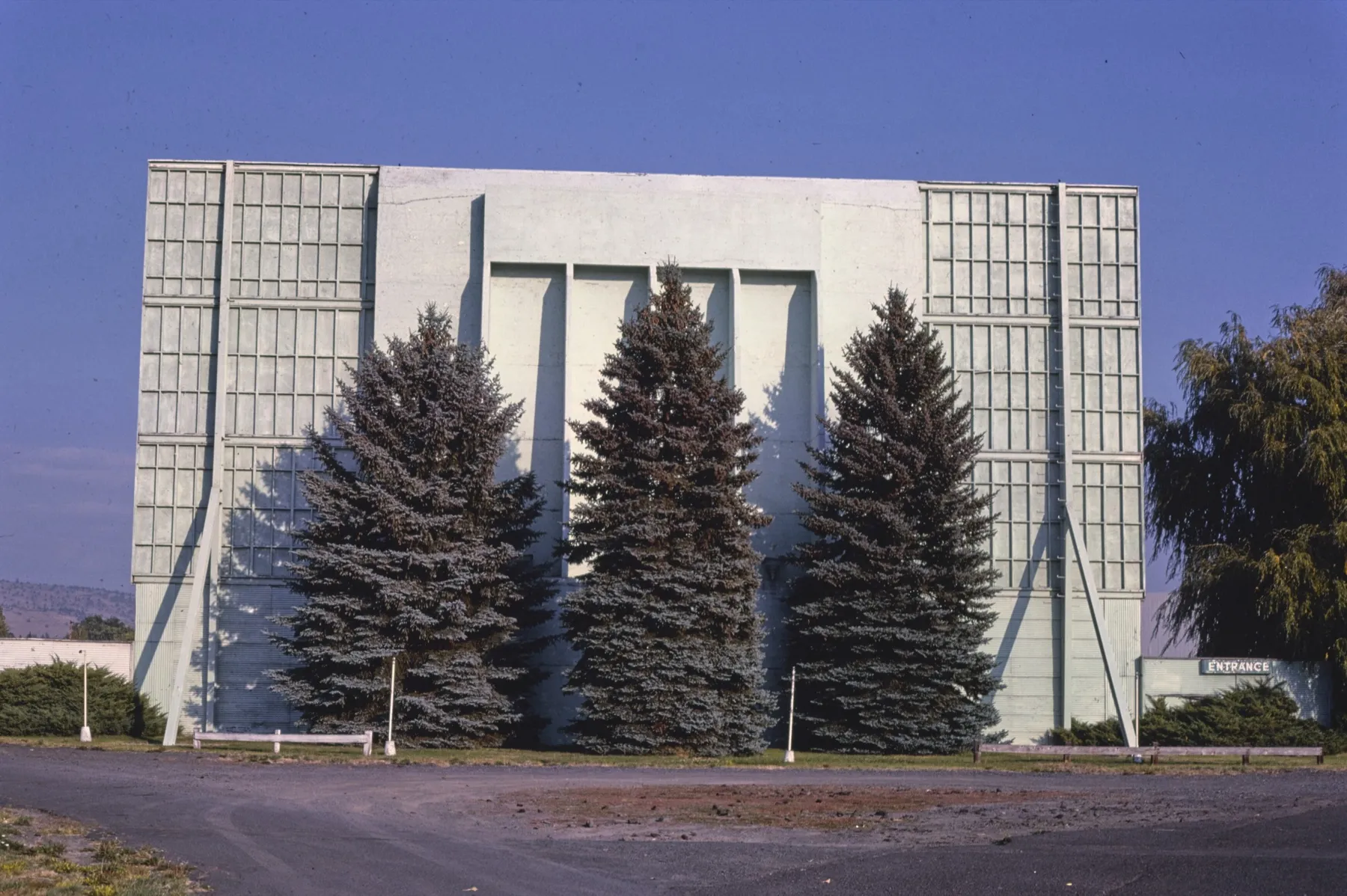 The concrete back of a drive-in screen tower with three tall blue spruces planted in front of it and a small Entrance sign at the right