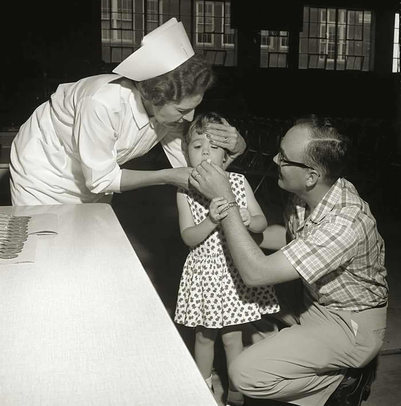 A nurse in a white cap steadies a little girl’s head while her father, kneeling, puts a sugar cube in her mouth beside a table lined with spoons